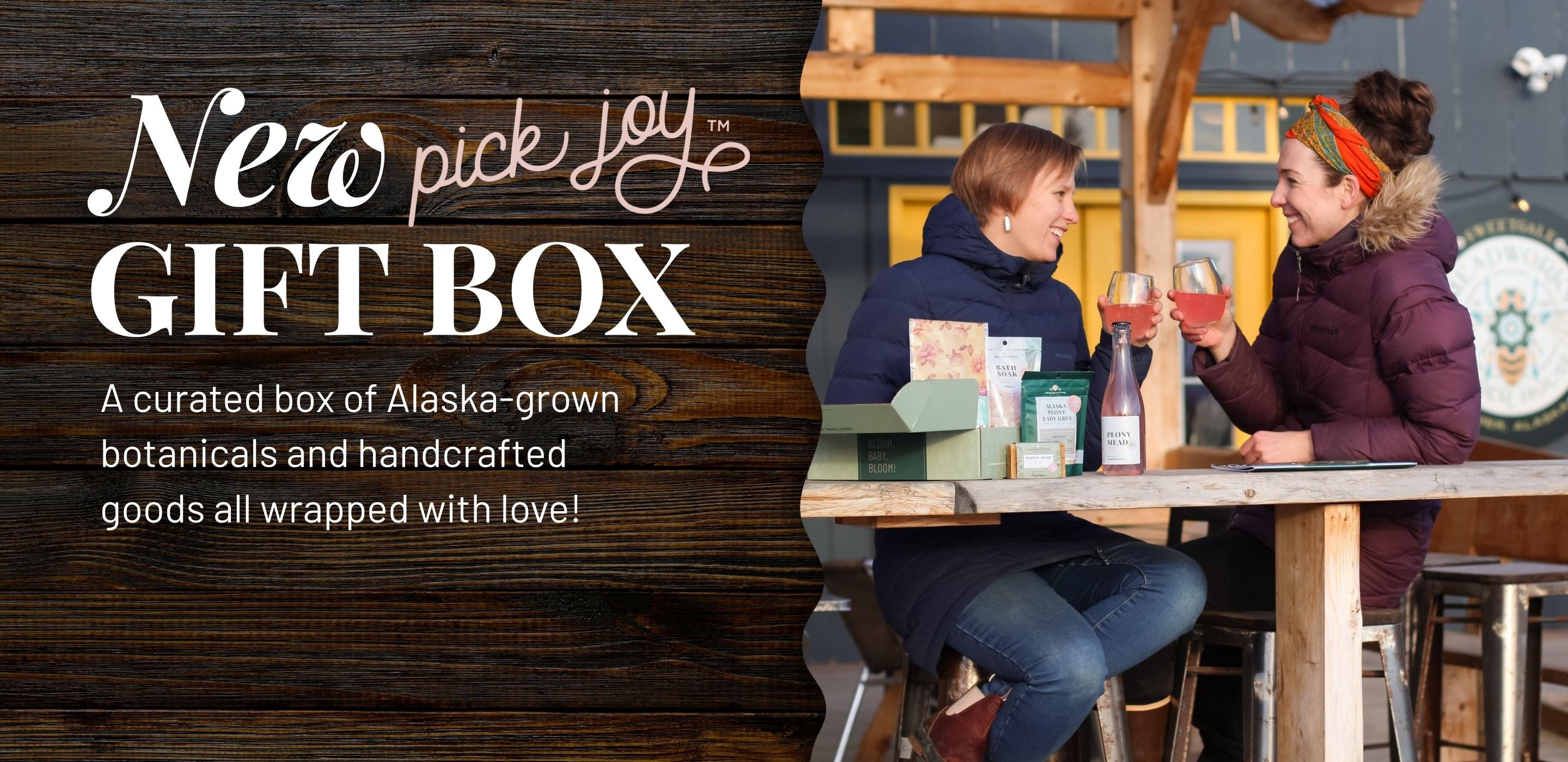 Two people sitting at a table with a 'New Pick Joy Gift Box' advertisement on a wooden background | Alaska Beauty Peony Cooperative | Homer, AK